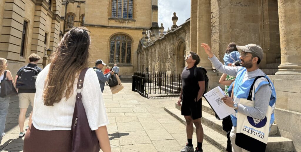 An Uncomfortable Oxford tour guide points at a historic college in Oxford while people look on.