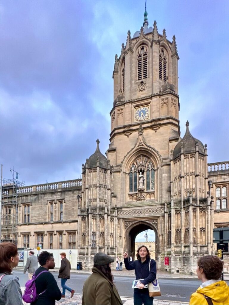 A group of tour attendees stand in front Christ Church College with an Uncomfortable Oxford guide and learn about Jewish history in Oxford.