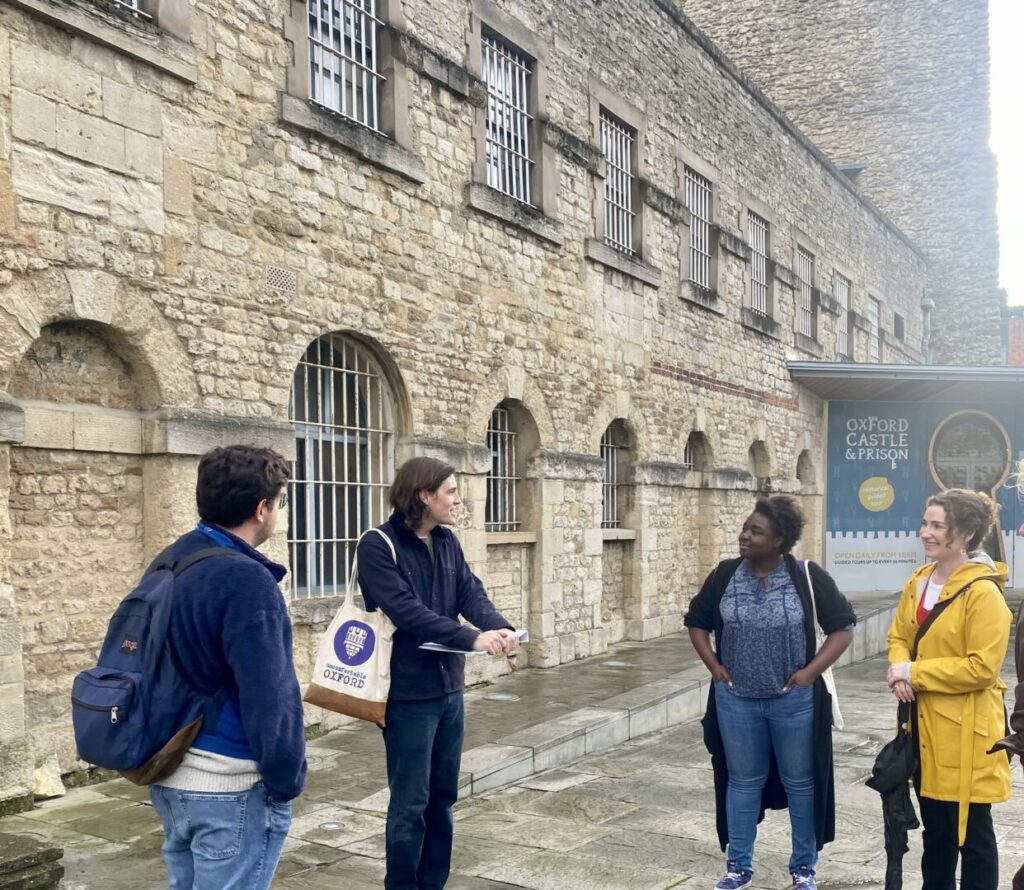 A group of tour attendees stand in front of an Uncomfortable Oxford guide and learn about medieval history in Oxford.