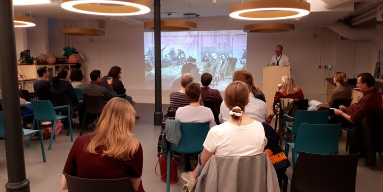 A man stands at the front of a lecture room and presents to an audience.