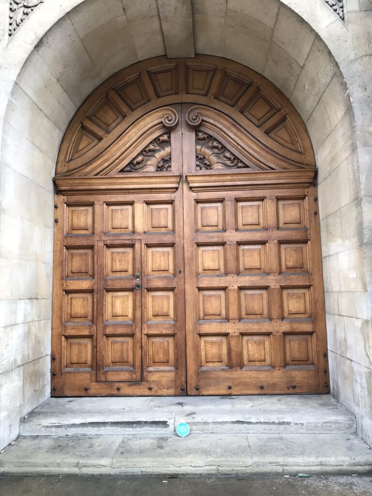 Large and old wooden door in a building, small blue plaque laid against the entrance steps.