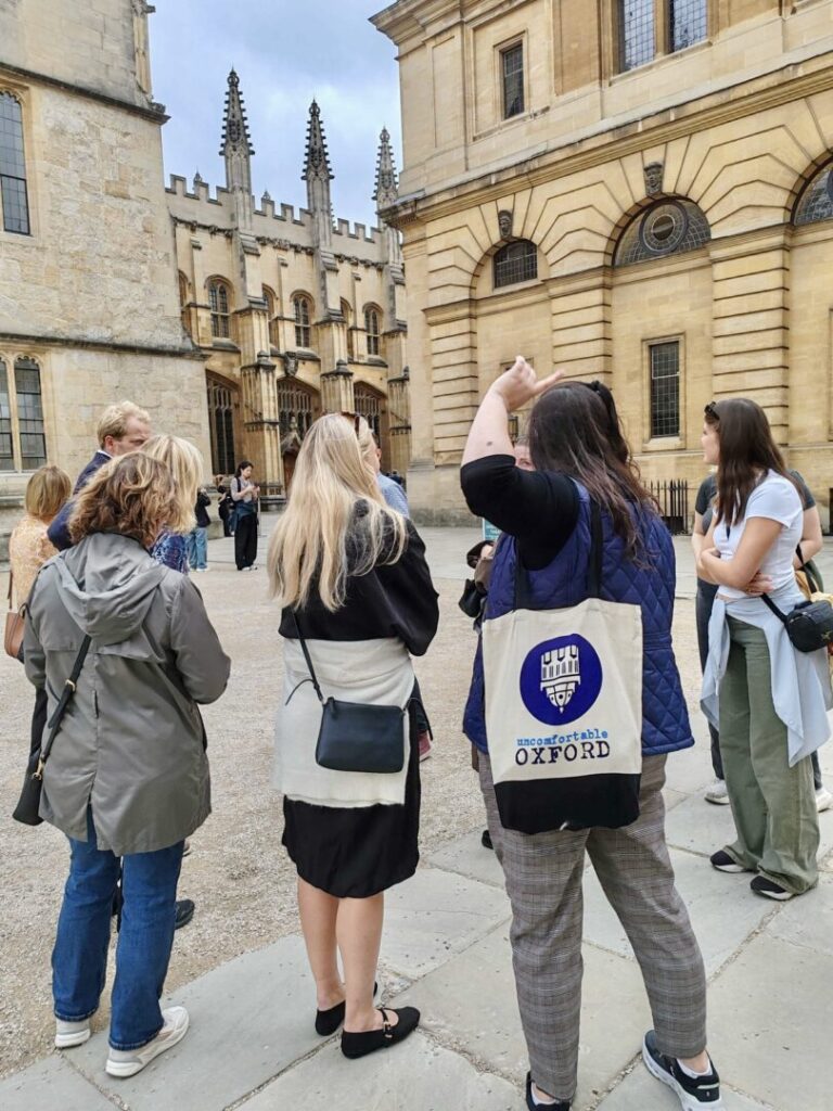 An Uncomfortable Oxford guide gestures at the Bodleian Library on a tour while a tour group looks on.